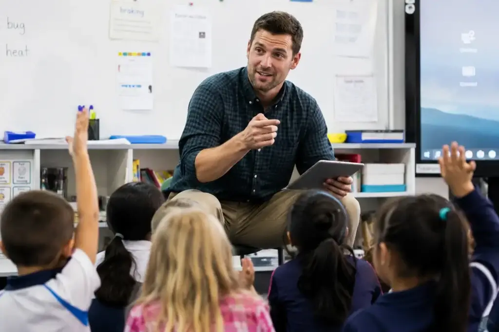 Primary school teacher holding a tablet and calling on students with their hands raised in the classroom.
